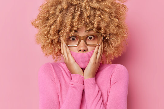 Portrait of shocked puzzled woman covers muth with collar of jumper cannot believe her eyes looks with terrified expression wears eyeglasses isolated over pink background hears surprising news
