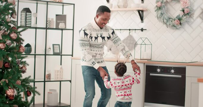 African American dad and little child listen to xmas music and dancing in modern home kitchen decorated with new year tree on Christmas Eve Happy family enjoy free time and having fun holidays concept