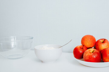 Red apples on plate, sugar, empty cup on white table. Front view of ingredients for making pie at home