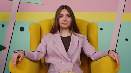Stylish bossy woman in purple jacket is sitting in yellow armchair in creative office space, looking at camera confidently, young and successful lady, Zoom in, Slow motion.