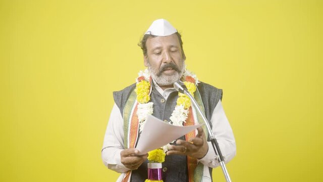 Politician seriously making oath ceremony with garland and white cap by holding papers in front of microphone on yellow background - concept of Politics, public speech or announcement