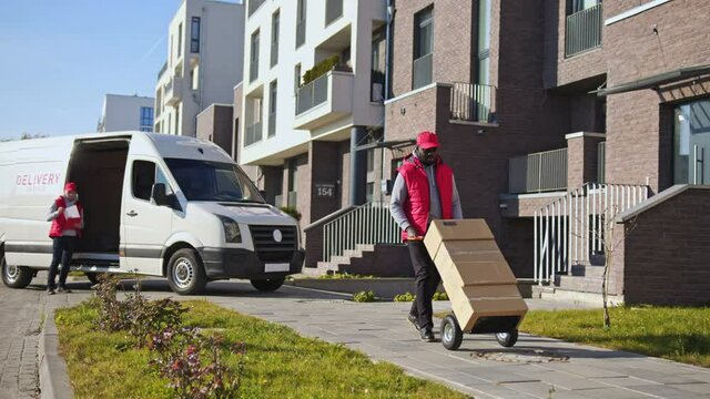 African american delivery man carring parcel boxes on a trolley from a van. Courier on the way to deliver postal parcel to a client.