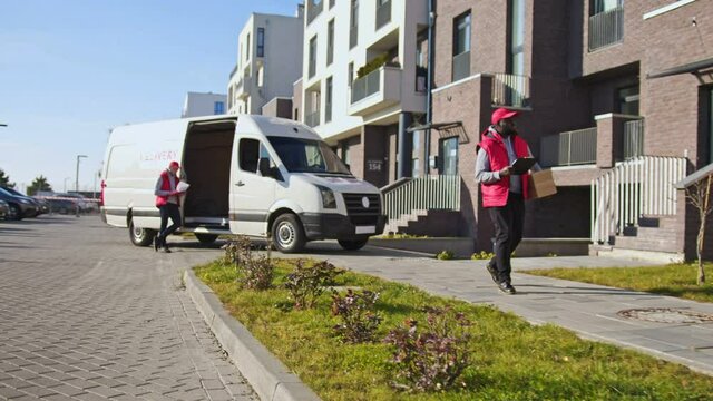 African american delivery man handling parcel box, walking along the street with tablet folder. Courier on the way to deliver postal parcel to a client.