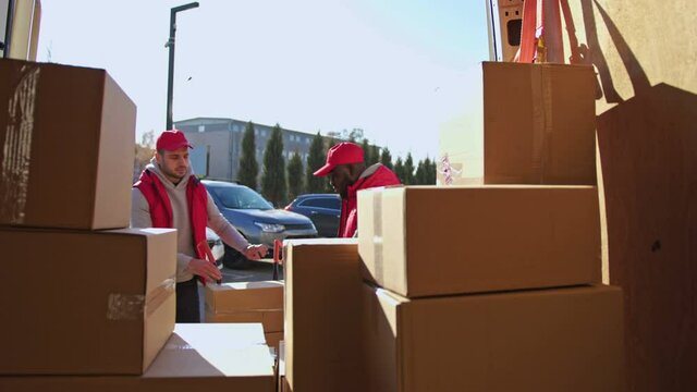 African american and caucasian couriers taking parcel boxes from a trolley to a van. Delivery men with a trolley on the way to deliver postal parcels to a client.