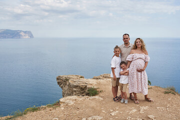 a family of four on the seaside stands against the backdrop of a very beautiful landscape. mom is pregnant, two sons and dad