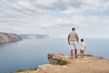 Fototapeta na wymiar young man and little son stand by the hand on the edge of the rocky coast with a beautiful sea view. back view
