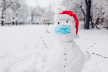 Snowman wears medical mask and Santa hat in snowy winter park. Safety protective measures during covid coronavirus