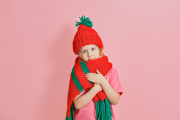 caucasian toddler girl in red knitted scarf and hat on pink background, christmas mood concept copy space