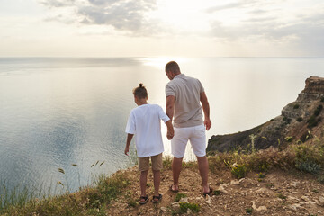 Fototapeta premium the boy holds his dad's hand and they look at the bottom of the cliff against the backdrop of a beautiful rocky sea coast going into the distance