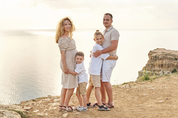 pregnant mom, dad and two sons. each son hugs a parent against the backdrop of a beautiful warm sunset sky and seascape.