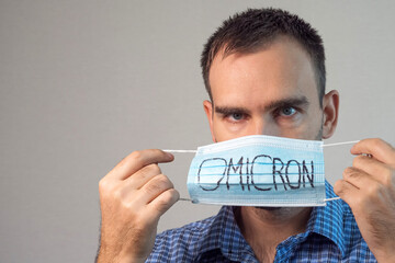 A man in a blue plaid shirt holds a blue medical mask with Omicron written on it in front of his...