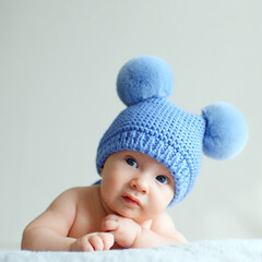 Sweet newborn baby in a blue hat with pompoms looks into the camera. A calm two-month-old infant is lying on a blanket. A beautiful, cute little child with blue eyes looks into the camera. .