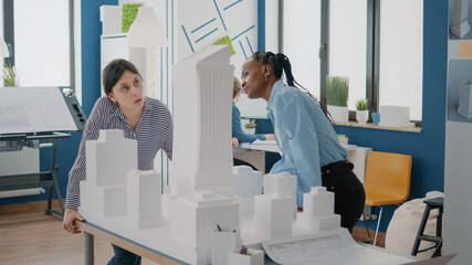 Team of multi ethnic women analyzing maquette on table to design building model. Workmates brainstorming ideas together to plan urban construction structure and architectural layout.