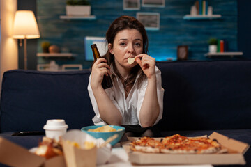 Sad woman drinking craft beer and eating potato chips from bowl at table with large pizza and...