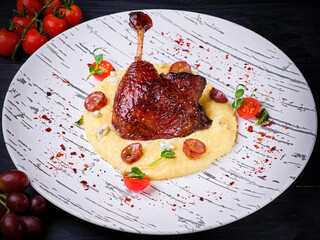 Traditional French Duck leg confit served on mashed potatoes with Dor blue cheese, grapes and Cherry tomatoes. Restaurant Main dish on white plate on black wooden background with ingredients
