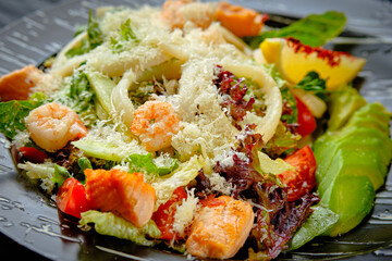 Spicy Seafood salad with grilled salmon, shrimp, squid, avocado, cherry tomatoes, lettuce served with parmesan cheese. Close up to Warm salad on black plate. Restaurant menu dish
