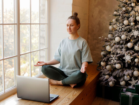 Portrait of young woman meditating while sitting near Christmas tree using laptop for online yoga lessons.