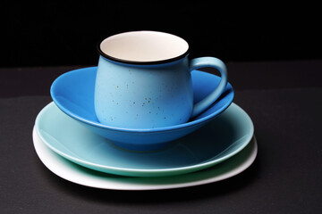 A ceramic mug in the shape of a blue pot and various plates stand in a stack on top of each other.