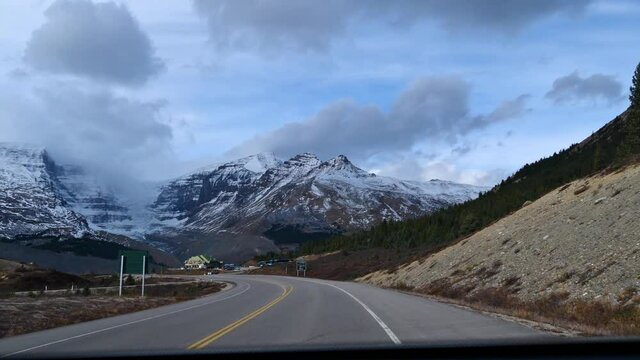 Passenger Front View Through Windshield Of Car Driving On Icefields Parkway In Jasper National Park, Alberta, Canada In The Rocky Mountains With Approaching Visitor Center Near Athabasca Glacier On Cl