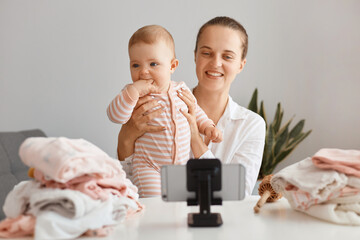 Indoor shot of smiling satisfied woman blogger sitting at table with toddler kid and recording video for her vlog, making content with her daughter, posing in front of phone on tripod.