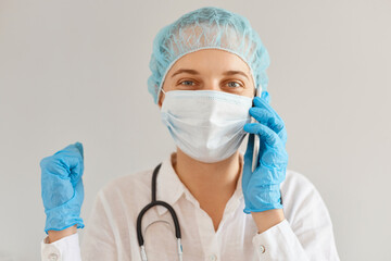 Happy positive young adult woman doctor wearing medical cap, surgical mask, gown and rubber gloves, being at work in hospital, talking phone, hearing good news, clenching fists.