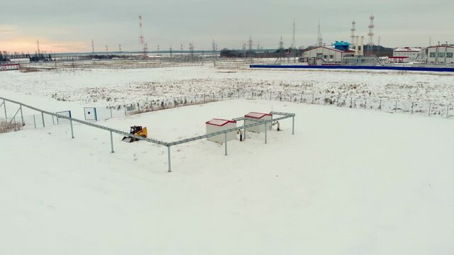 A drone flies over an industrial area in a snowy area, a mini-loader tractor clears the snow from the service area. Oil and gas industry in winter.