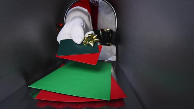 Santa Claus placing a pair of gift cards with a gold bow into a rural mailbox on top of a green and red envelopes, then giving a thumbs up to the camera, viewed from inside the mailbox.
