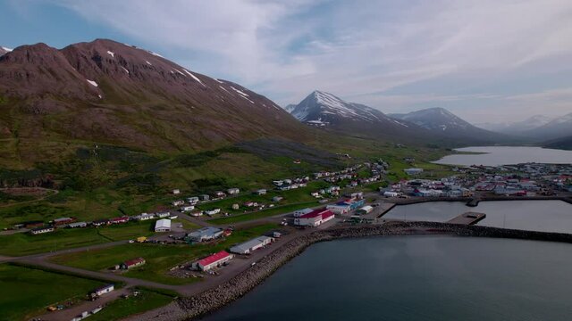 Aerial shot turning to reveal town in Icelandic fjord with snowy mountains in distance. Coastal village.