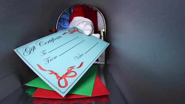 Santa Claus placing a gift certificate into a rural mailbox on top of a green and red envelopes, then giving a thumbs up to the camera, viewed from inside the mailbox.