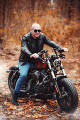 Fototapeta na wymiar Bald brutal man in sunglasses and a leather jacket sits on a motorcycle on the road in the autumn forest