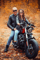 Fototapeta na wymiar Cute couple near a red motorcycle in the autumn forest. Relationship concept. A pair of bikers in leather jackets.