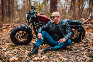 Brutal model man in sunglasses and a fashionable leather jacket in biker style sits near a motorcycle in the autumn forest