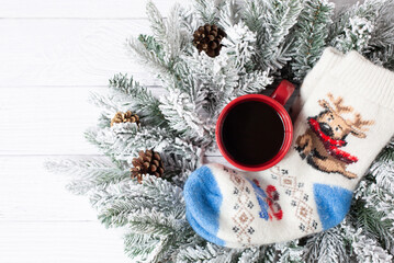 Warm woolen knitted socks with a New Year's pattern, a red cup with black coffee and spruce snow branches are on a white wooden background. Free space for text. View from the top point.