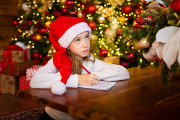 Fototapeta na wymiar Merry Christmas and Happy Holidays. little child girl in santa claus cap writes letter Santa Claus and dreams of a gift background New Year tree indoors. Greeting card, Christmas mood concept.