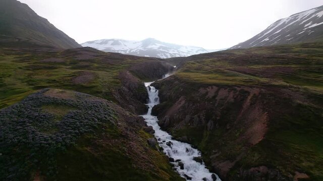 Low aerial shot with creek and small waterfalls below in lush Icelandic landscape. Beautiful scenic view and snowy mountains in distance.