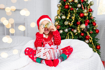 a child girl with Christmas balls on her ears at the Christmas tree in a red sweater and Santa Claus hat on New Year's Eve in a white bed smiling, fooling around and laughing with a gift