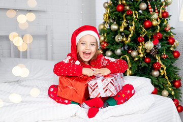 a child girl with gifts at the Christmas tree in a red sweater and Santa Claus hat on New Year's Eve or Christmas at home on a white bed screams with happiness