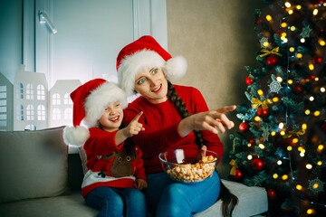 Fototapeta na wymiar A young mother and her daughter watch movies at home on Christmas day pointing at the screen
