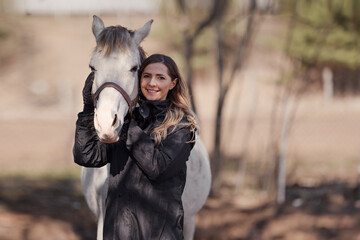 Young woman standing next to white Arabian horse smiling, sun shines on blurred trees and field background