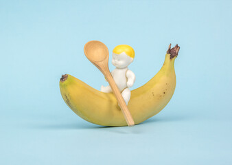 A figurine of a baby sitting on a banana with a wooden spoon. The concept of healthy baby food.
