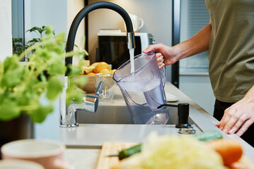 Woman pouring water from faucet into water filter jug at the kitchen