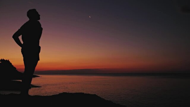 Silhouette of a man looking at the Moon and stars over sea ocean horizon.	
