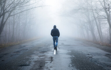 Lonely man walking in fog away road. Rural landscape with road in morning mist. Warm autumn colors....