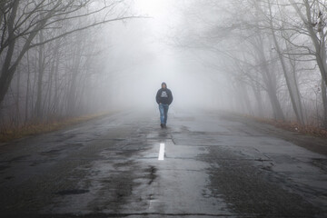 Lonely man walking in fog away road. Rural landscape with road in morning mist. Warm autumn colors. Dark mysterious background