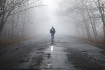 Lonely man walking in fog away road. Rural landscape with road in morning mist. Warm autumn colors....