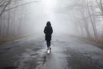Lonely woman walking in fog away road. Rural landscape with road in morning mist. Warm autumn...