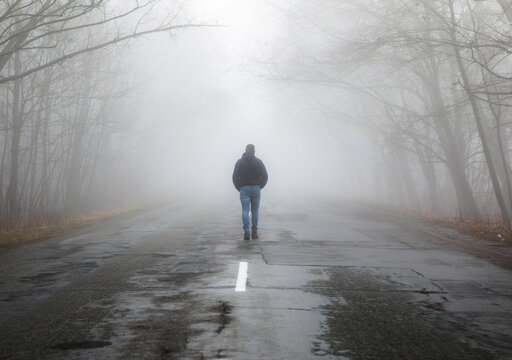Lonely man walking in fog away road. Rural landscape with road in morning mist. Warm autumn colors. Dark mysterious background