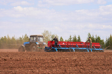 Obraz premium Tractor with a trailed seeder in the field. There are two farmers on the seeder who monitor the process of sowing seeds and fertilizing from the hopper.