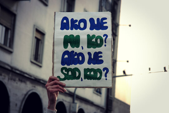 A woman holds a cardboard poster in hand at a protest on the street in Serbia. On it written, if not we, who, if not now, when?