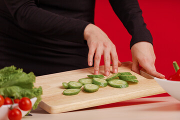 Female hands cut fresh cucumber on a wooden board on the table with a large cook knife on a red background - Powered by Adobe